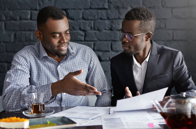 Attractive African businessman in glasses and suit holding papers, showing presentation of his project to cheerful business partner, who smiling happily, supporting his idea with positive gesture